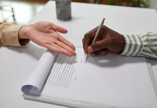 an man writing his signature on a contract while his attorney is showing him where to sign